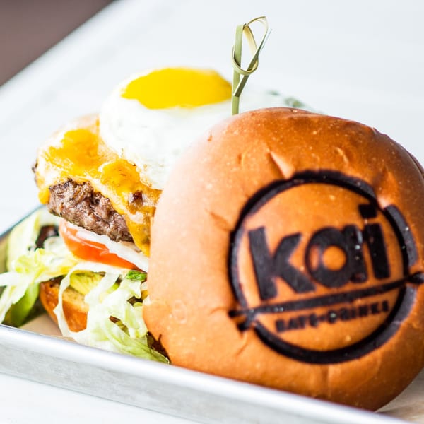 A Kai Eats + Drinks burger with melted cheese, fried egg, lettuce, and tomato on a metal tray.