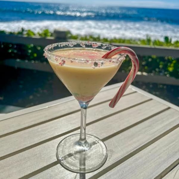 A festive cocktail with a candy cane garnish sits on a table overlooking the ocean.