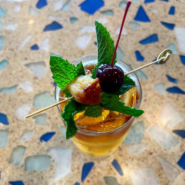 Kai Eats + Drinks cocktail with mint, cherry, and candied bacon on a terrazzo table.