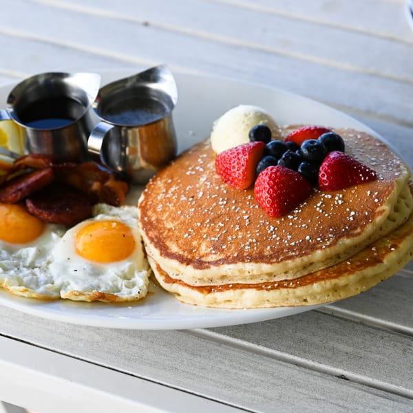 Pancakes with berries, ice cream, fried eggs, bacon, and syrup on a white plate.