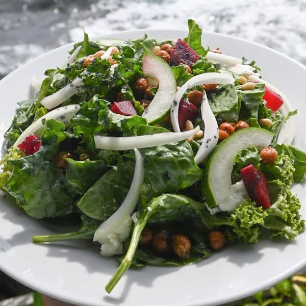 A vibrant green salad with sliced cucumbers, red beets, chickpeas, and pine nuts on a white plate.