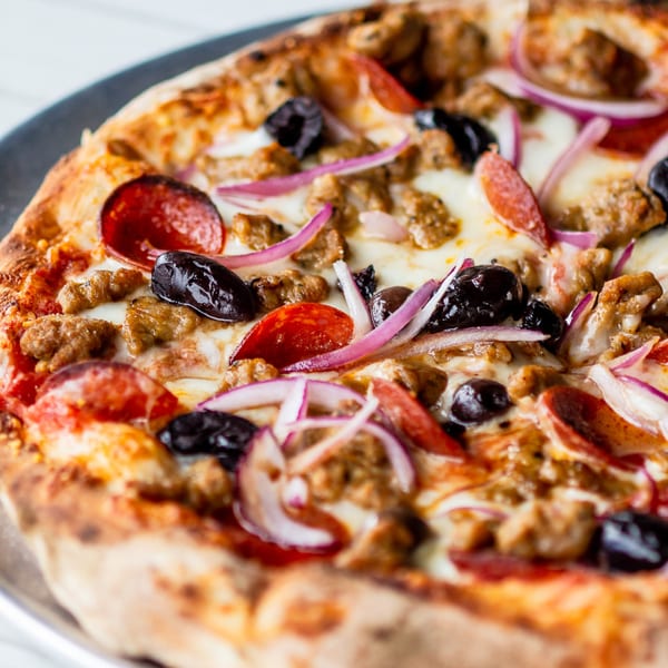 Close-up of a Kai Eats + Drinks pizza with pepperoni, sausage, black olives, and red onions.